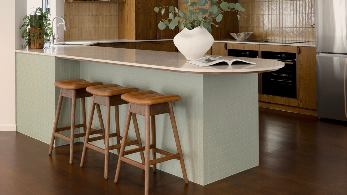 A modern kitchen island with a pale sage green textured base, a light beige stone countertop, and three wooden bar stools. The background shows a kitchen with wood cabinets, a tiled backsplash, and stainless steel appliances.